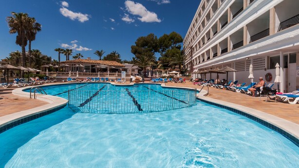 Una amplia piscina exterior con múltiples carriles, rodeada de tumbonas y sombrillas, y un edificio con balcones al fondo.