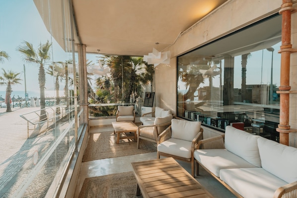 Un área de descanso moderna con mobiliario de mimbre, un amplio ventanal y vistas a palmeras y la playa.