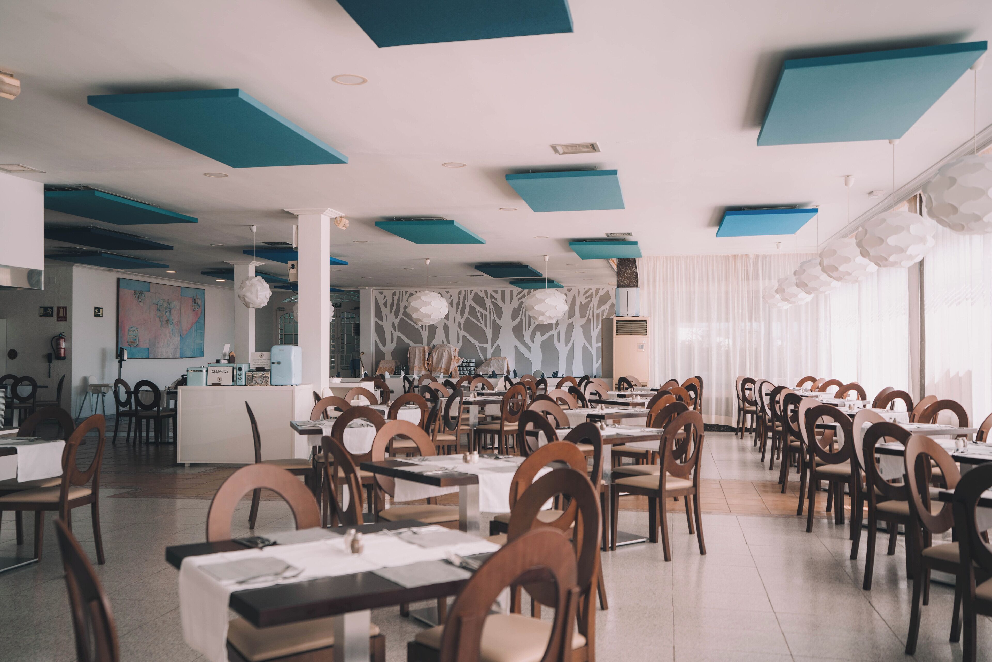 A spacious restaurant with high ceilings, teal accents, and a modern design. Tables are neatly arranged with chairs, and there is a counter in the background.
