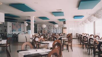 A spacious restaurant with high ceilings, teal accents, and a modern design. Tables are neatly arranged with chairs, and there is a counter in the background.