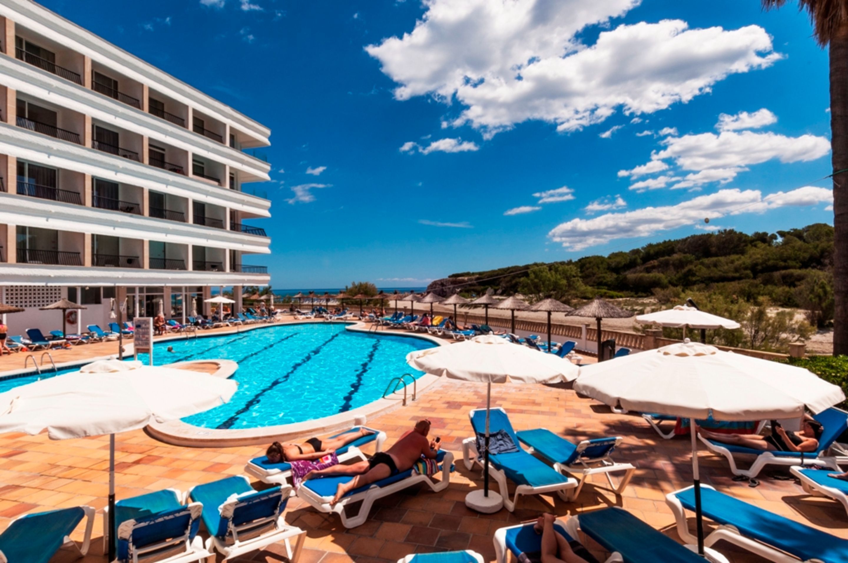 A pool area with lounge chairs and umbrellas, a building with balconies, and a clear blue sky.