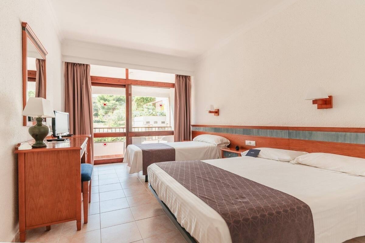 Een hotelkamer met twee bedden, een bureau met lamp, een groot raam met gordijnen en uitzicht op groen buiten.