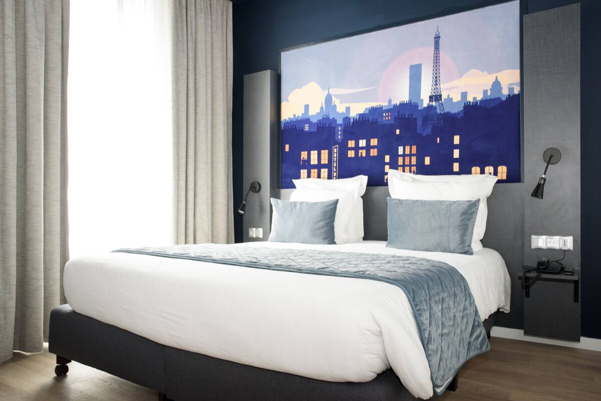 Une chambre d’hôtel moderne avec un grand lit, une œuvre d’art représentant un paysage urbain accrochée au mur, une table de chevet avec une lampe et une prise de courant.