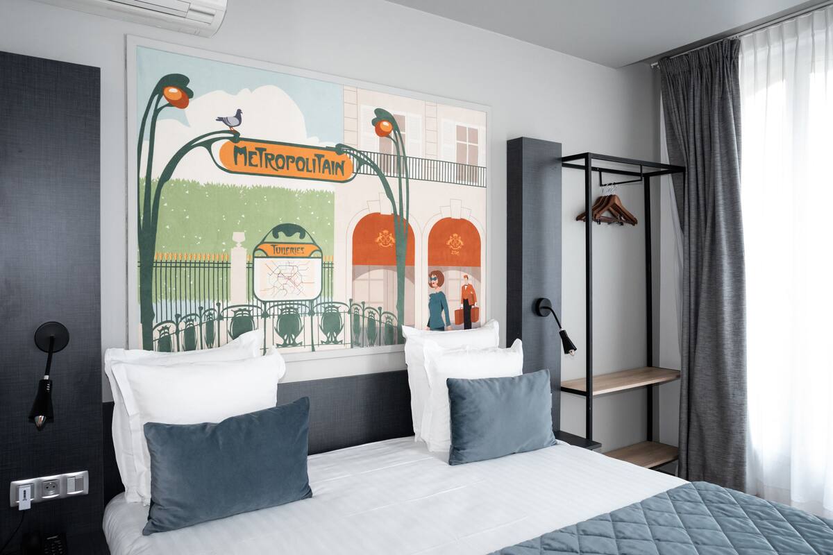 Une chambre d’hôtel comprenant un lit, deux oreillers, une table de chevet, une étagère fixée au mur et une fenêtre avec des rideaux.