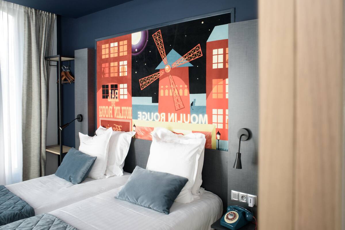 Une chambre d’hôtel avec deux lits, une grande fenêtre avec des rideaux, une fresque murale et un téléphone ancien posé sur la table de chevet.