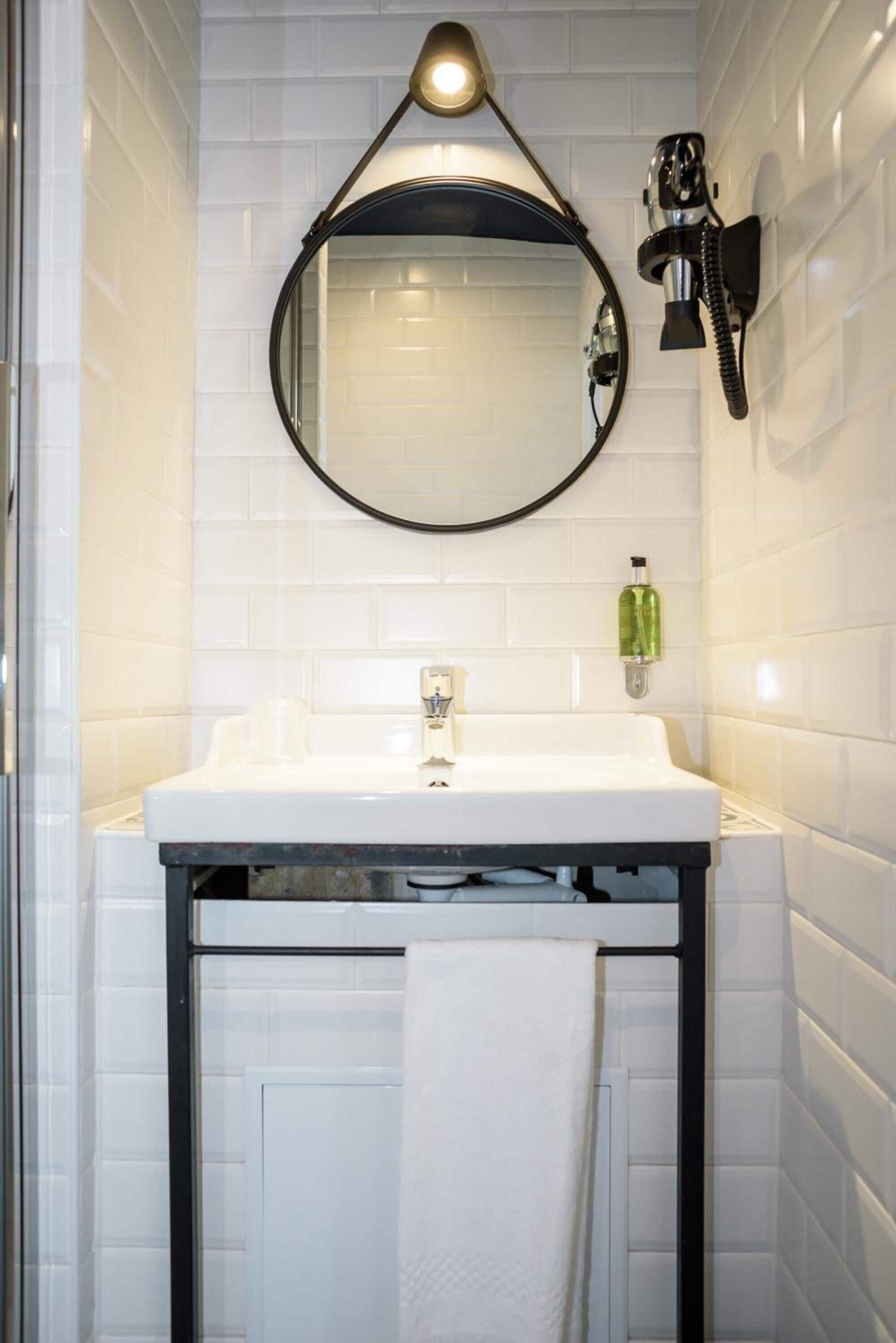 A bathroom with a white tiled wall, a white sink, a black-framed mirror, a wall-mounted telephone, and a green bottle of hand soap.
