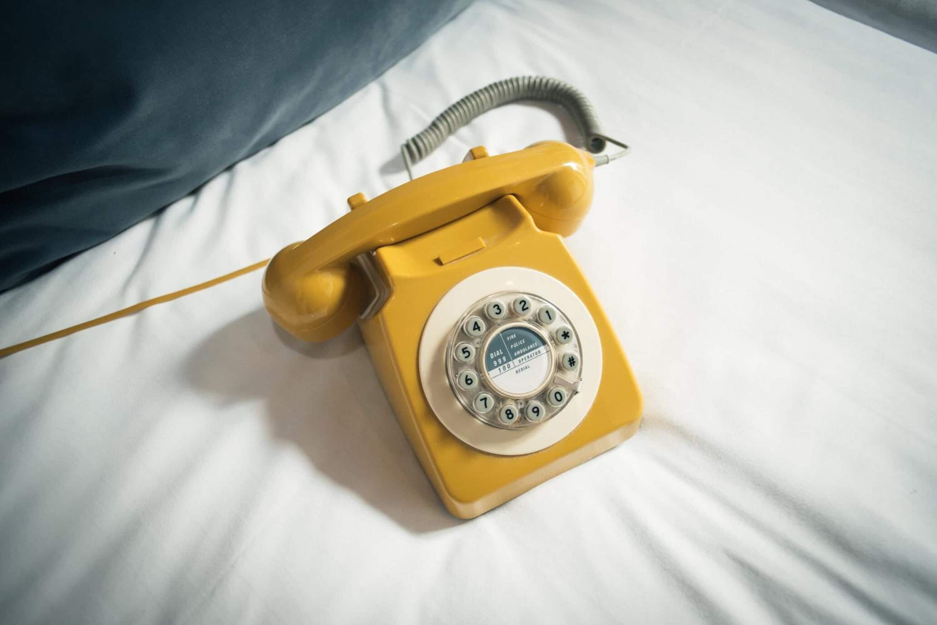Een gele telefoon met draaischijf op een bed met witte lakens.