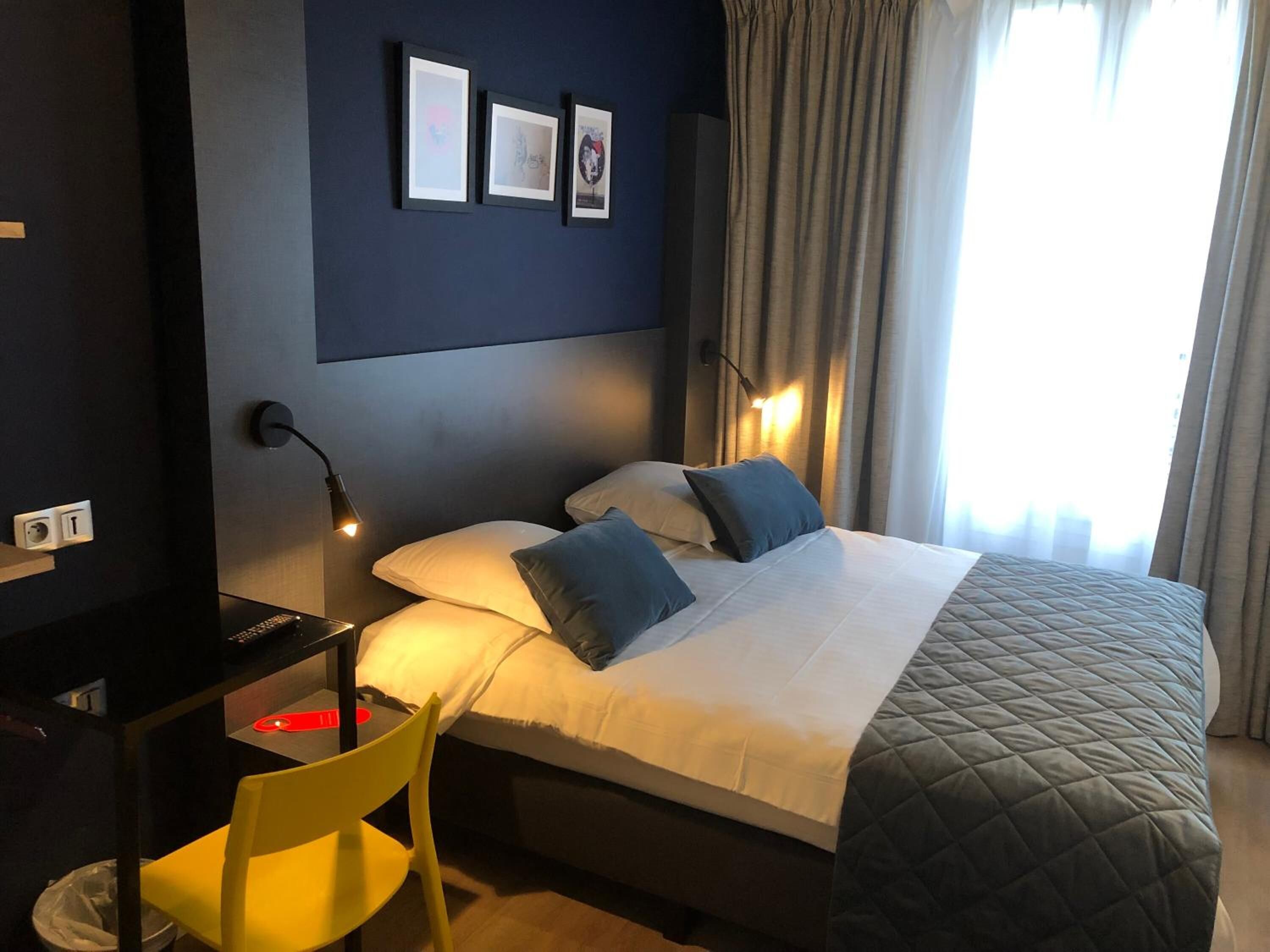 A hotel room with a bed, a desk with a chair, and a wall-mounted lamp.