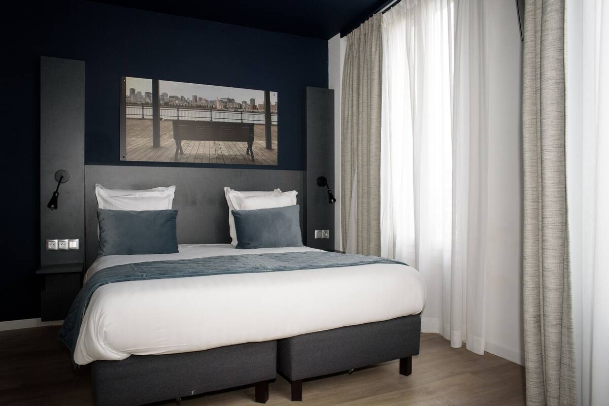 Une chambre avec un grand lit, un tableau représentant un paysage urbain accroché au mur et une vue sur un pont.