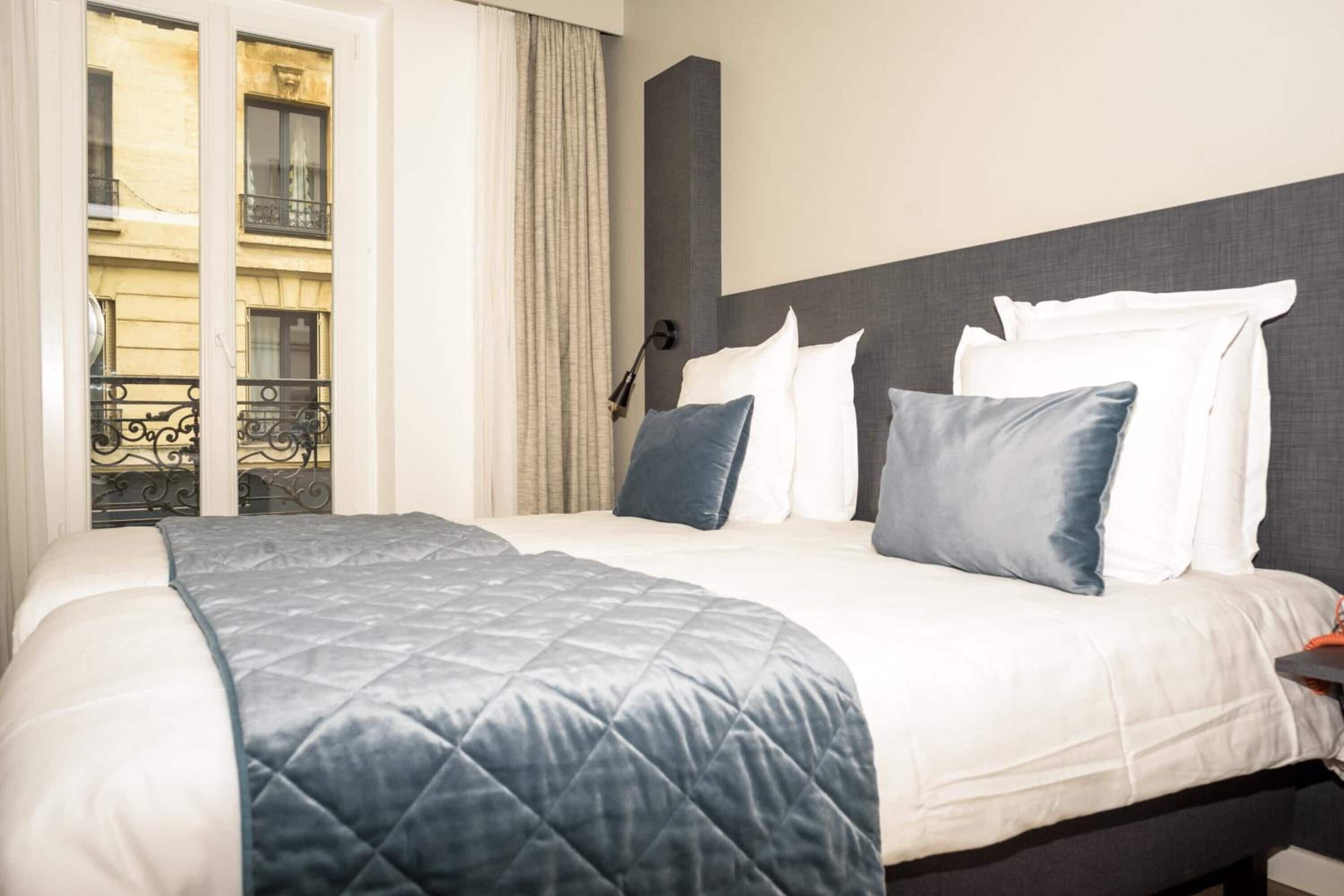 A hotel room with a bed, a headboard, a window with curtains, and a view of a building outside.