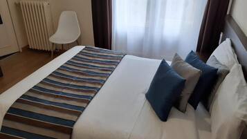 A hotel room with a bed, a chair, a radiator, and a window with curtains.