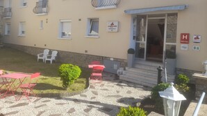 Un hotel con fachada color beige, múltiples ventanas con contraventanas, una zona de estar exterior roja y un pequeño jardín.