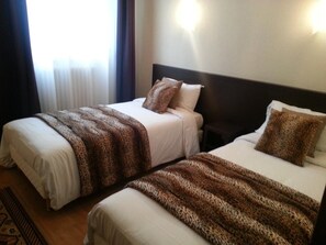 Dos camas individuales con cabeceras y almohadas a juego, una ventana con cortinas, y un aplique de pared.