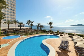 A swimming pool with lounge chairs and umbrellas, a building with balconies, and a view of the sea.