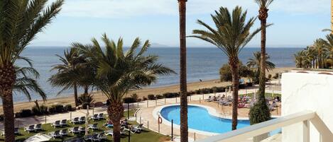 Zona de piscina en un resort, con palmeras, sillones de descanso y vista al mar.