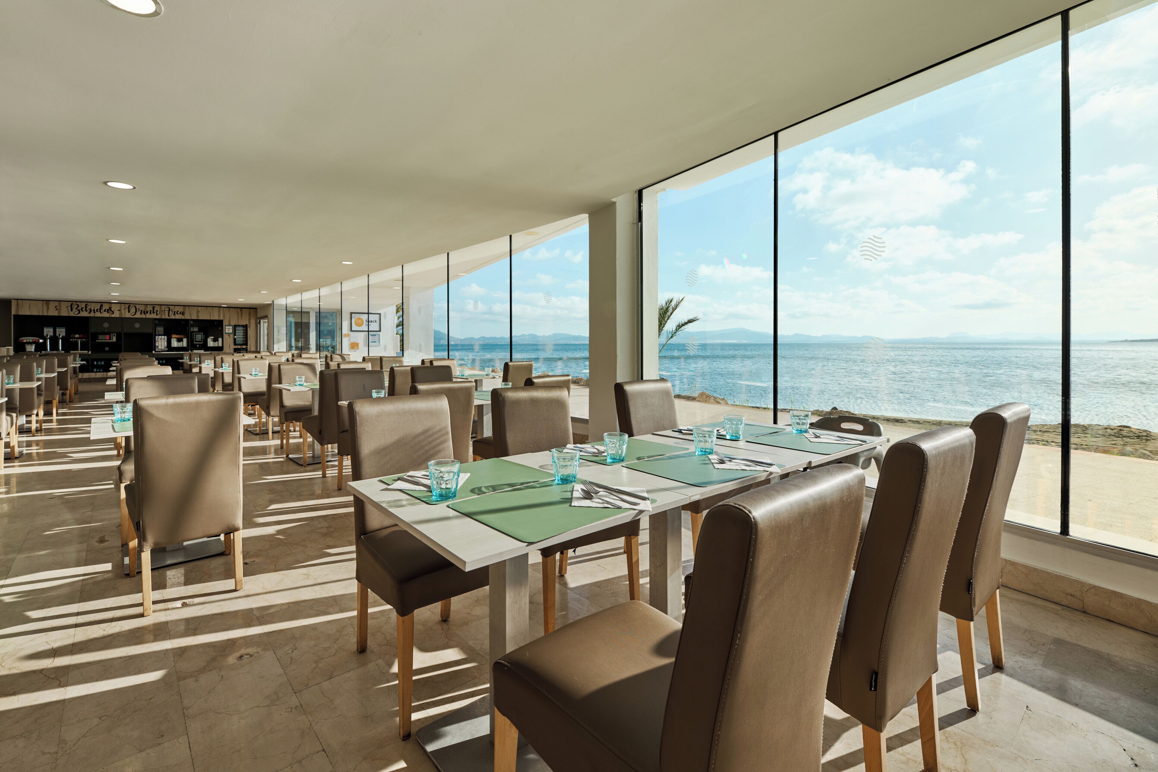 A spacious dining area with tables and chairs, large windows with a view of the sea, and a bar area in the background.