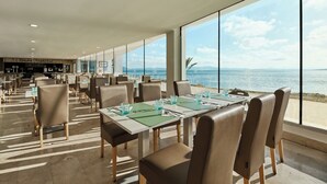 Una àmplia zona de dinar amb taules i cadires, grans finestres amb vista al mar, i una zona de bar al fons.
