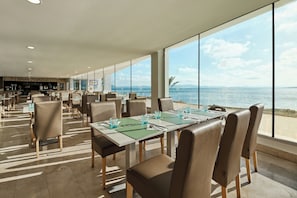 A spacious dining area with tables and chairs, large windows with a view of the sea, and a bar area in the background. - Aluasun Doblemar (San Javier)