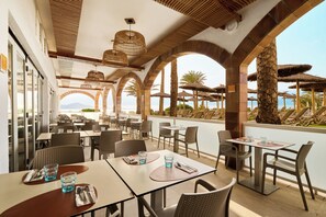 A restaurant with outdoor seating, tables with chairs, and a view of the sea.