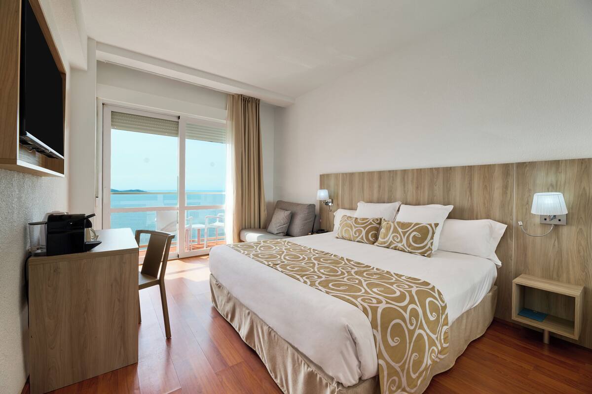 A hotel room with a bed, a desk, a chair, a nightstand, and a view of the sea.