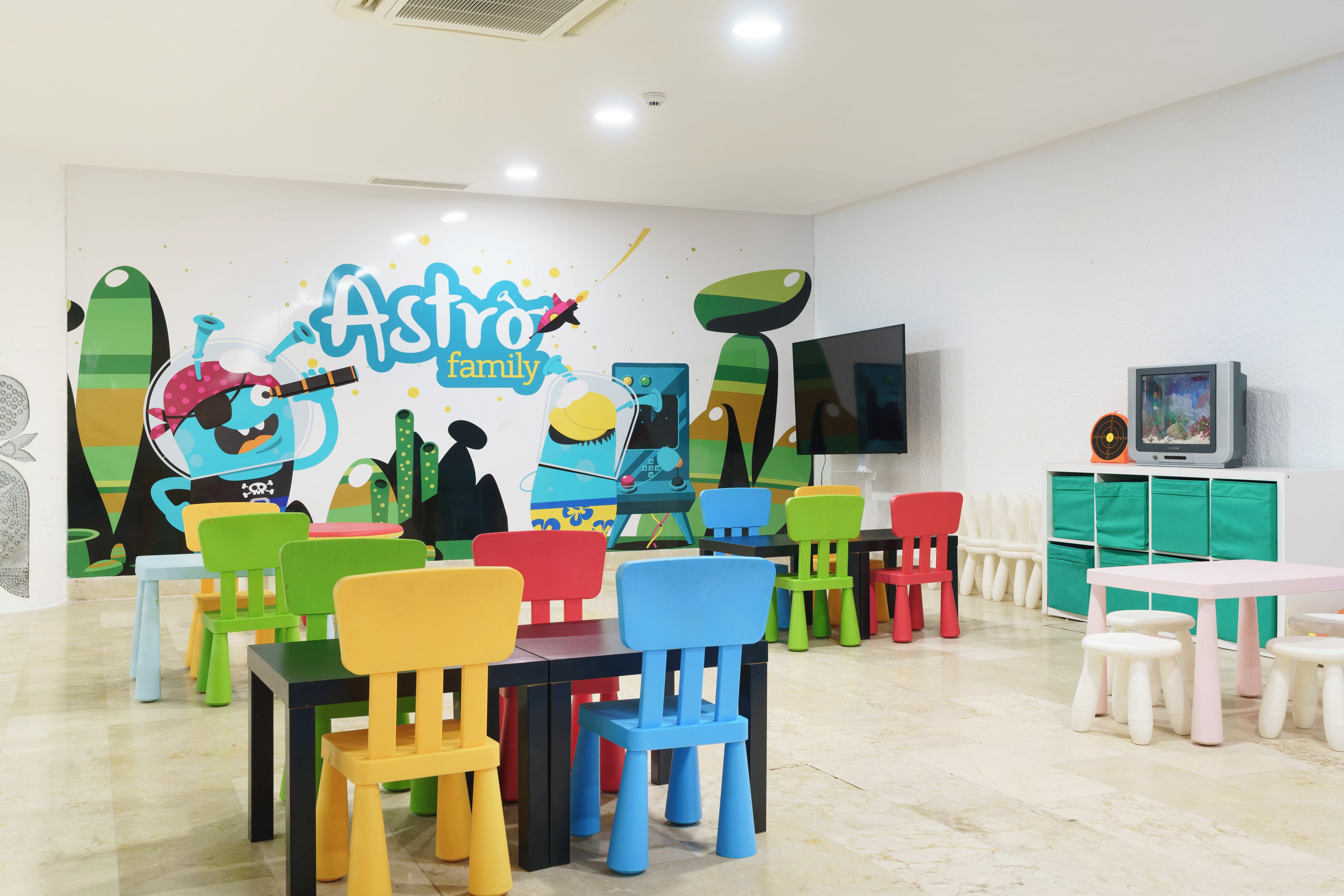A brightly colored children's playroom with a mural, multiple tables and chairs, and a television.
