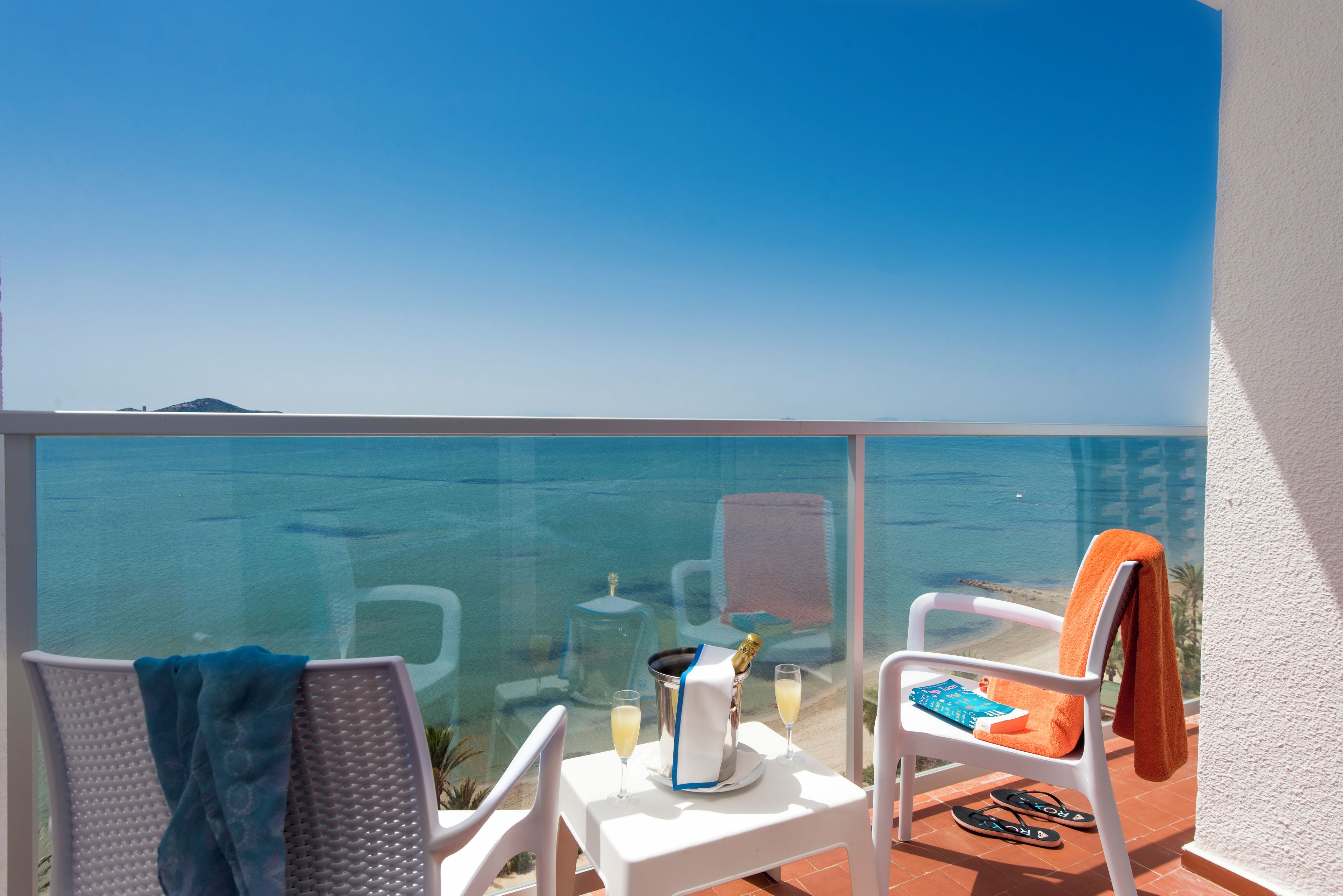 Un balcón con vista despejada al mar, dos sillas, una mesa con una botella y vasos, y una toalla colgada en una silla.
