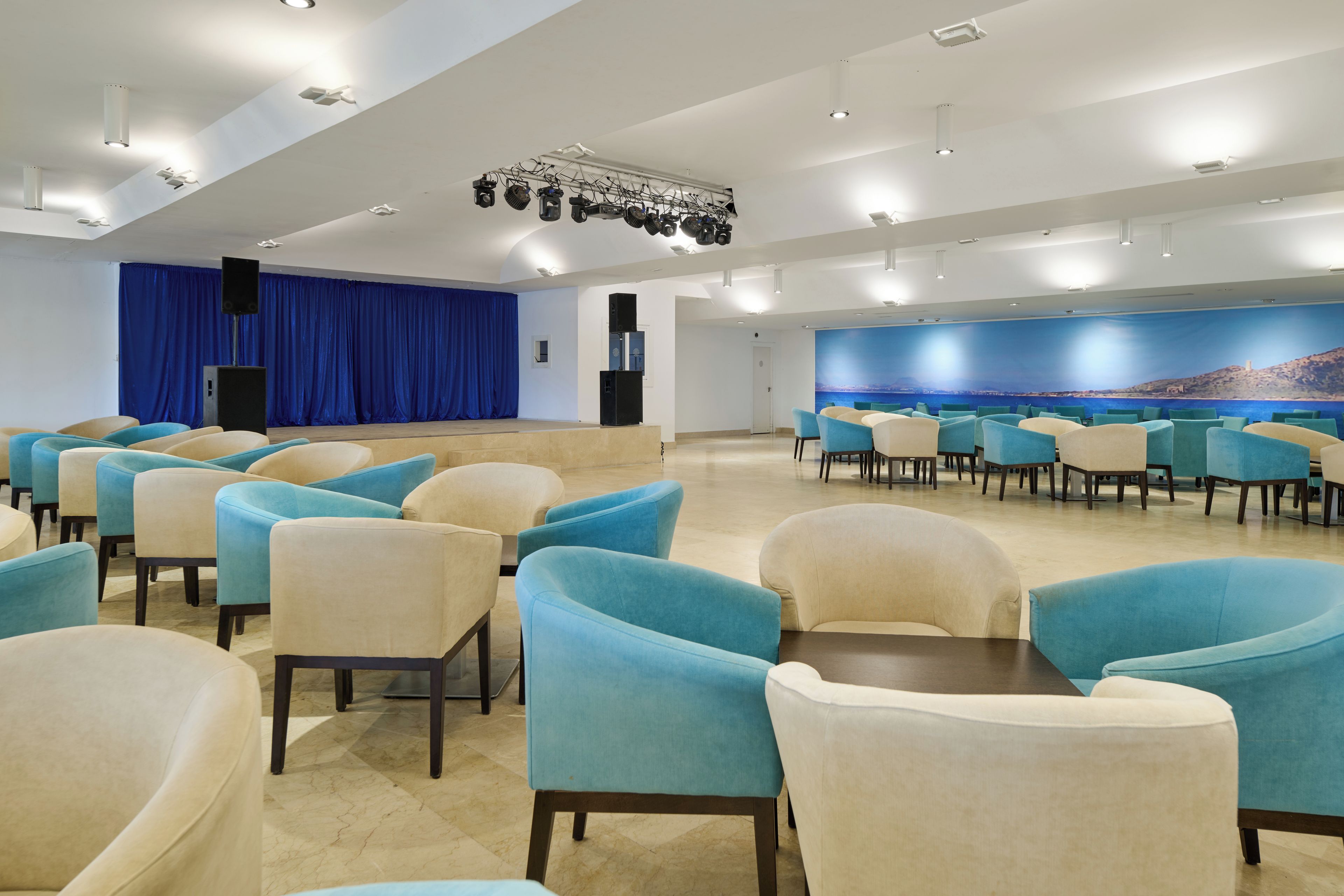 Een ruime congreszaal met rijen stoelen, een podium met een blauw gordijn en een groot muurschilderij.