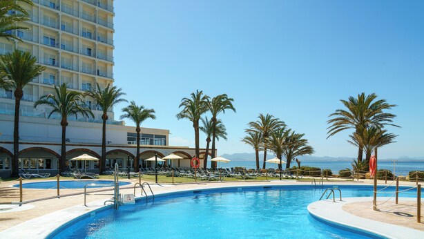 A swimming pool with palm trees and lounge chairs, a building with balconies, and a view of the ocean. - Aluasun Doblemar (San Javier)