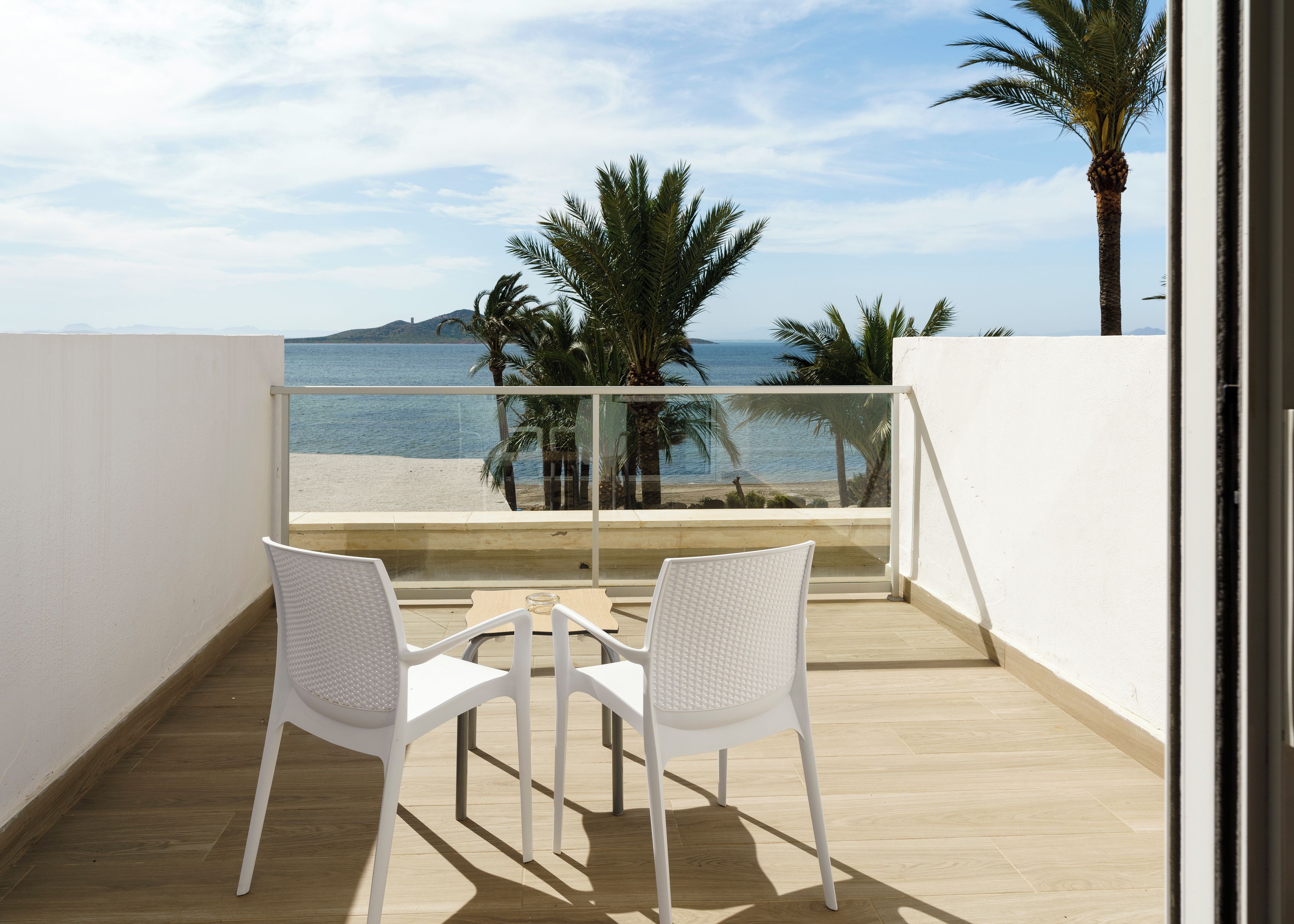Un balcone con due sedie bianche e un tavolino, vista su una spiaggia con palme e il mare.