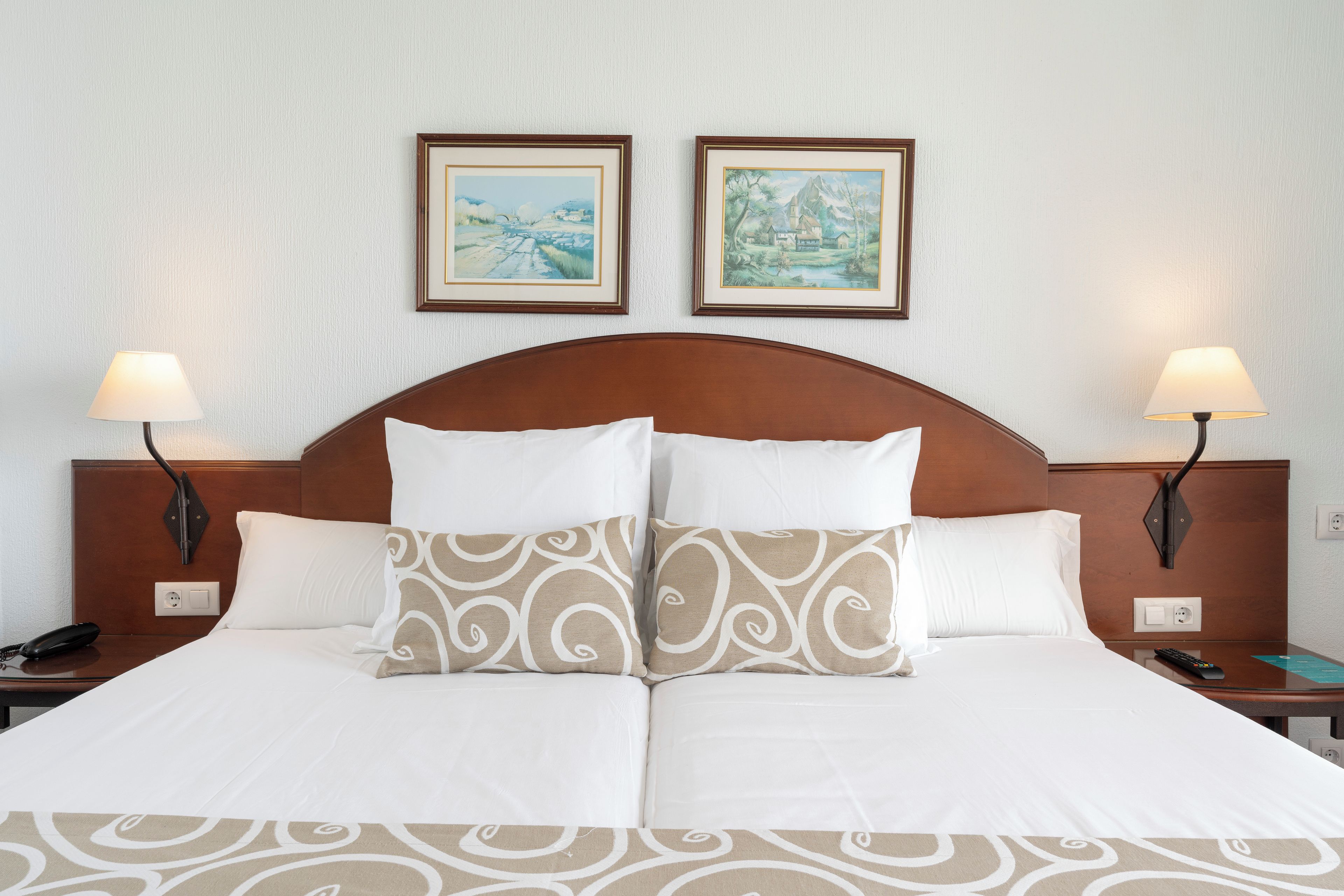 A double bed with white and patterned pillows, two bedside tables with lamps, and two framed paintings on the wall.
