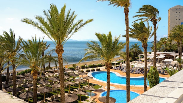 Un complex turístic amb piscina, palmeres i vistes al mar.