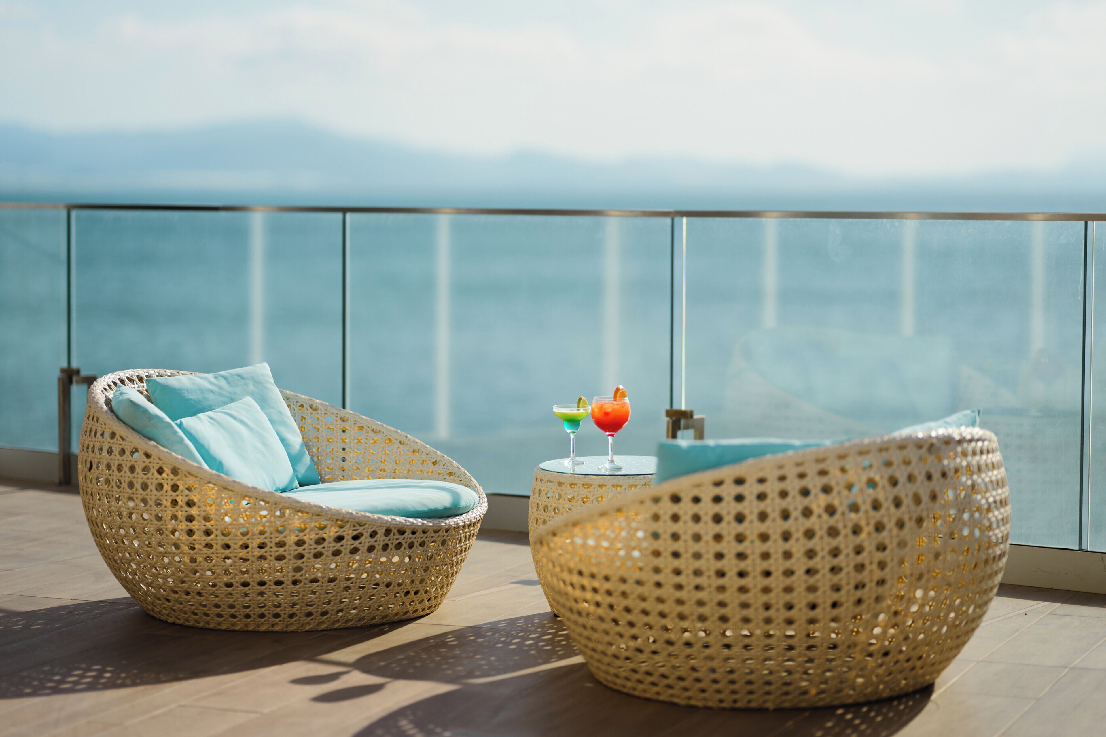 Wicker lounge chairs with cushions, a small table with a drink, and a view of the sea.