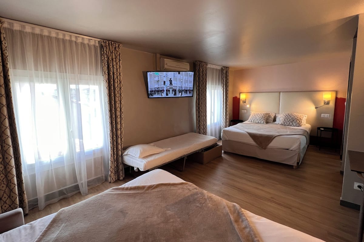 A hotel room with two beds, a TV mounted on the wall, and a window with sheer curtains.