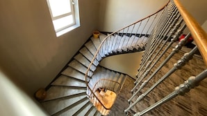 Una escalera de caracol con barandilla de metal y pasamanos de madera, que conduce a una zona de descanso con un ventanal pequeño y un cubo.