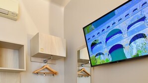 Habitación de hotel con un televisor montado en la pared, una mesita con dos tazas y una tetera, un perchero y una estantería con una radio.