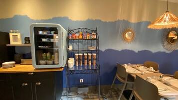 A dining area with a table set for two, a refrigerator, and a shelf with jars.