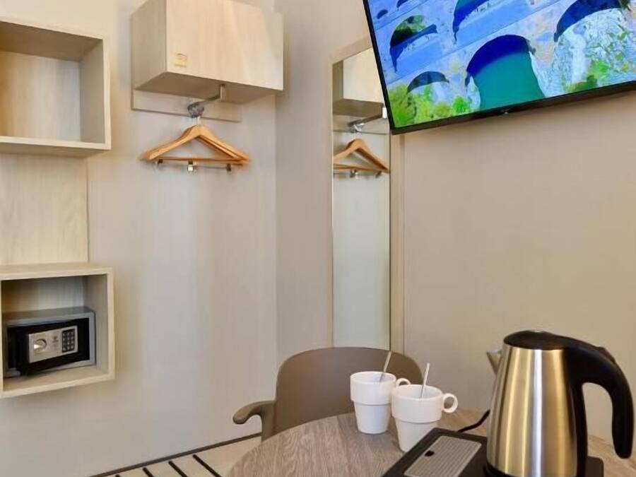A hotel room with a round table, a kettle, a coffee maker, a TV on the wall, a mirror, and a small safe.
