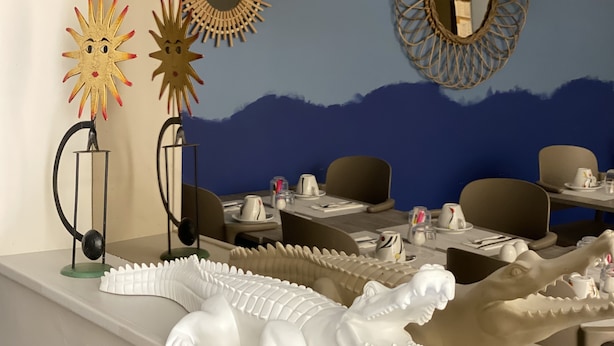 Un comedor con mesas dispuestas para el servicio, decoración mural y una gran escultura blanca de cocodrilo en la encimera.