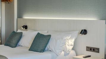 A neatly made bed with white linens and two green pillows, a bedside table with a remote control, and a wall-mounted light fixture.