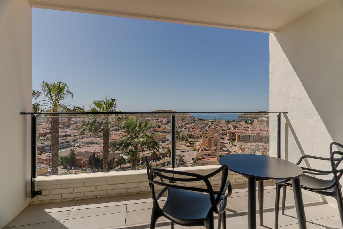 A balcony with a table and chairs, overlooking a coastal cityscape with palm trees and buildings.