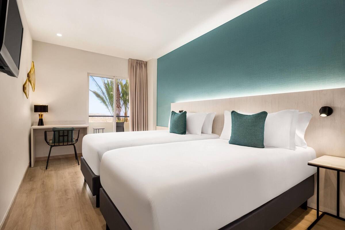 A modern hotel room with two beds, a desk, and a view of palm trees.