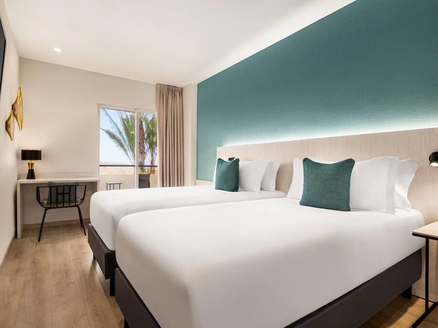 Moderni hotellihuone, jossa on kaksi sänkyä, työpöytä ja näkymä palmuille.