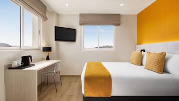 Habitación de hotel con cama, escritorio, silla, televisor en la pared y ventanas amplias.