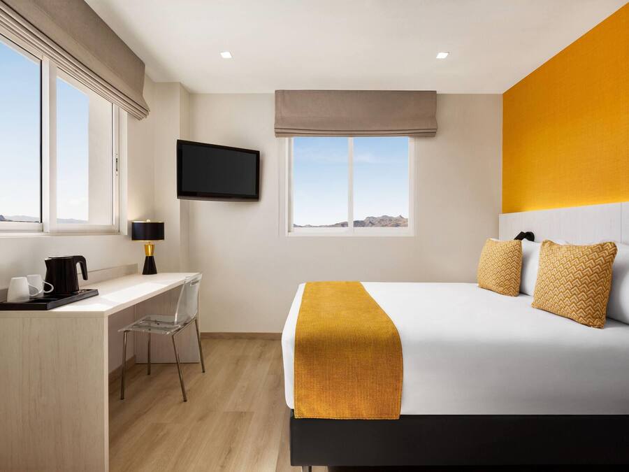 Habitació d'hotel amb llit, escriptori, cadira, televisió penjada a la paret i grans finestres.
