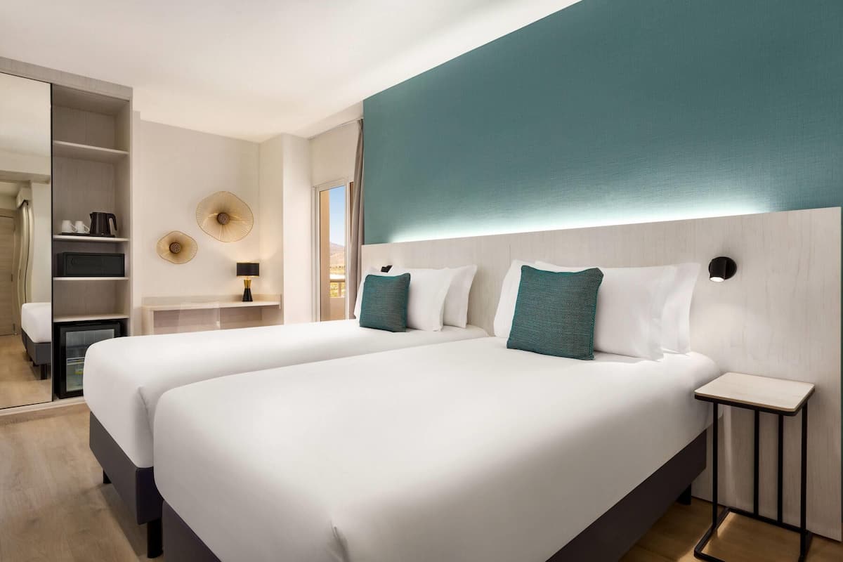 A hotel room with two beds, a small table, a shelf with toiletries, and a view of the outdoors.