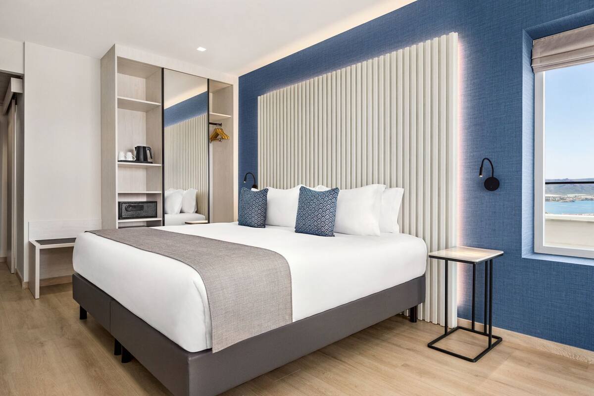 A modern bedroom with a large bed, a bedside table, a wardrobe, and a window with a view.