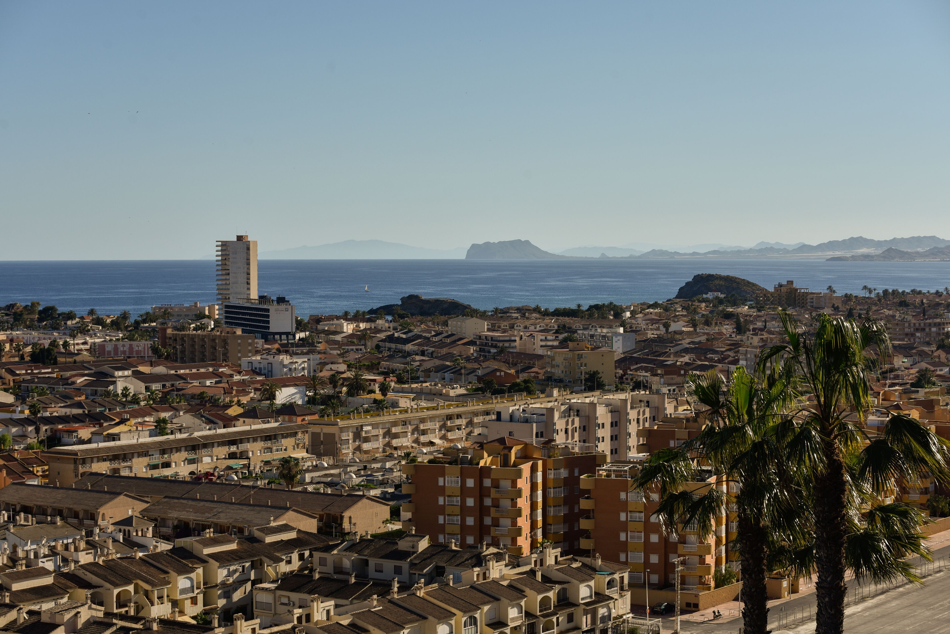 Un paisaje urbano costero con una mezcla de edificios residenciales y comerciales, un rascacielos destacado y una vista despejada del mar y montañas lejanas.
