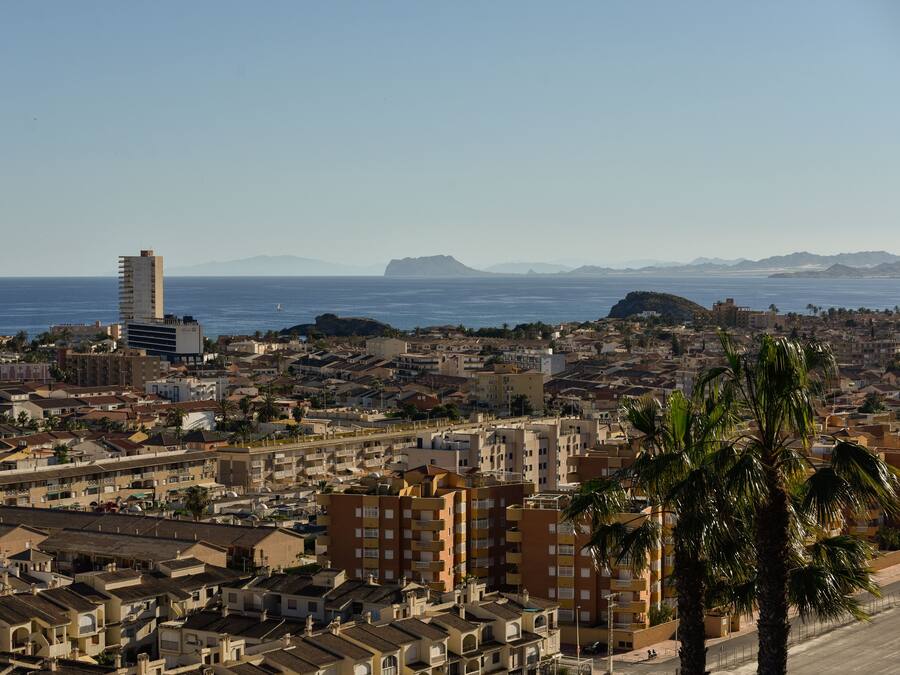 Un paisaje urbano costero con una mezcla de edificios residenciales y comerciales, un rascacielos destacado y una vista despejada del mar y montañas lejanas.
