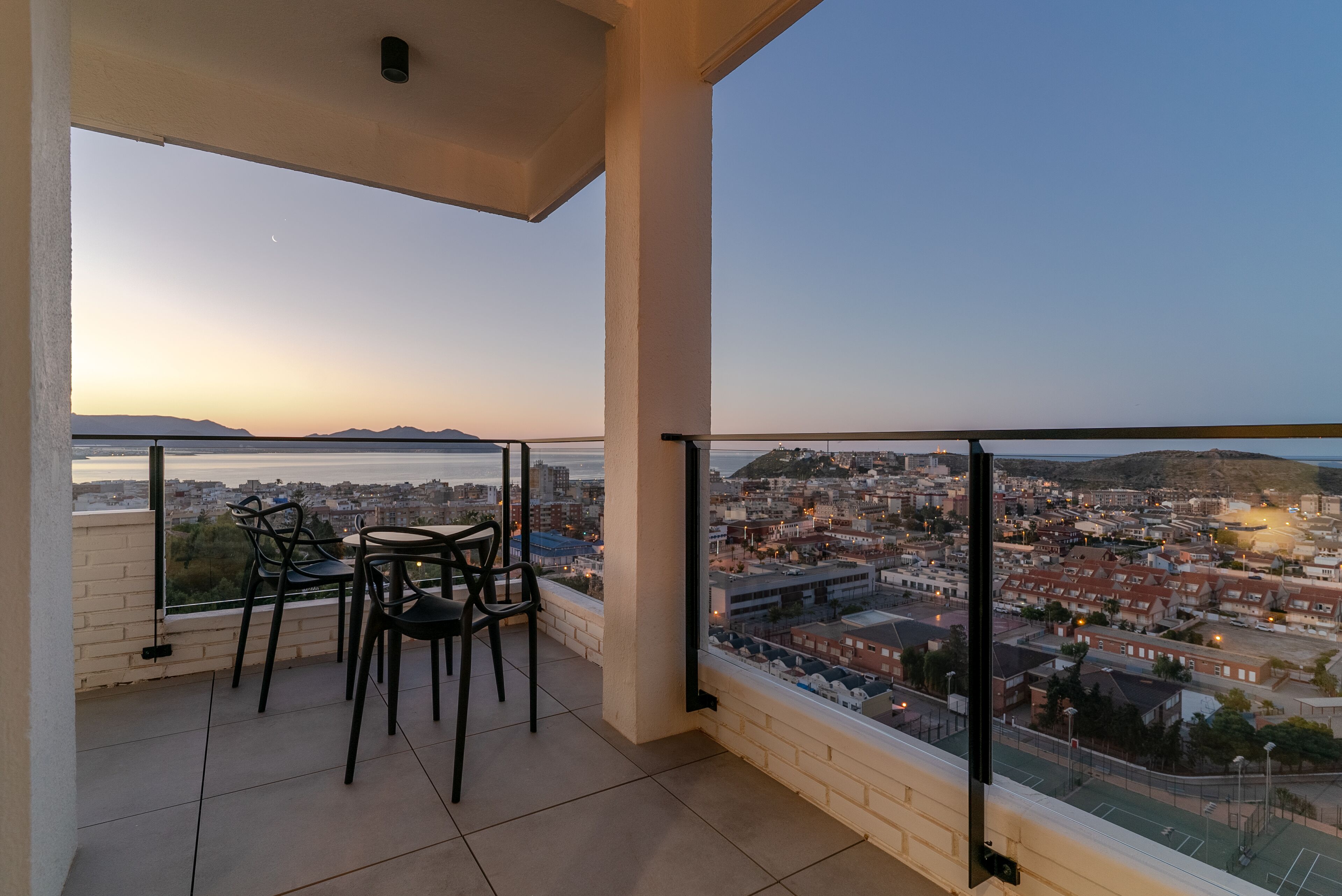 Un balcón con mesa y sillas, con vista a un paisaje urbano y una masa de agua al atardecer.