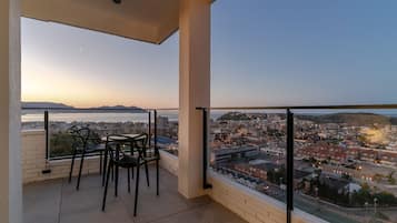 Un balcon avec une table et des chaises, offrant une vue sur le paysage urbain et une étendue d’eau au coucher du soleil.